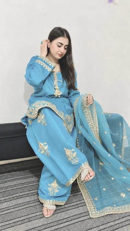 Seated model showcasing a sky blue Farshi Shalwar suit with embroidered wide trousers and matching dupatta.